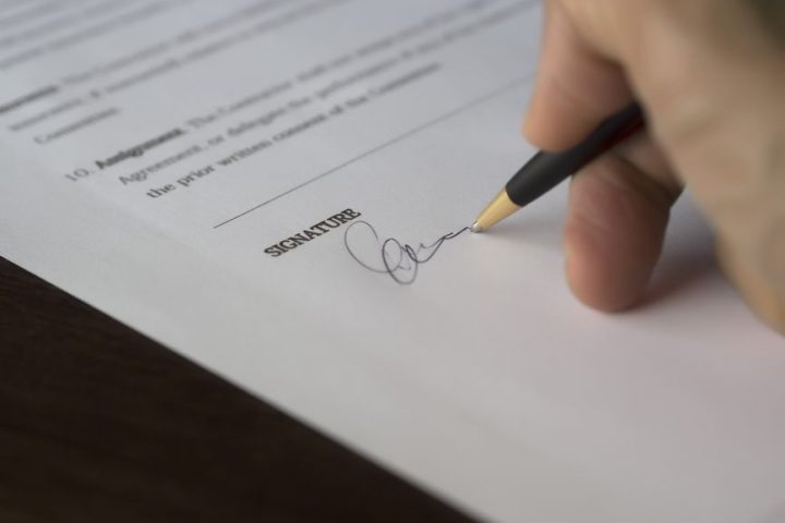 A hand signs a formal contract with a pen on a wooden desk.